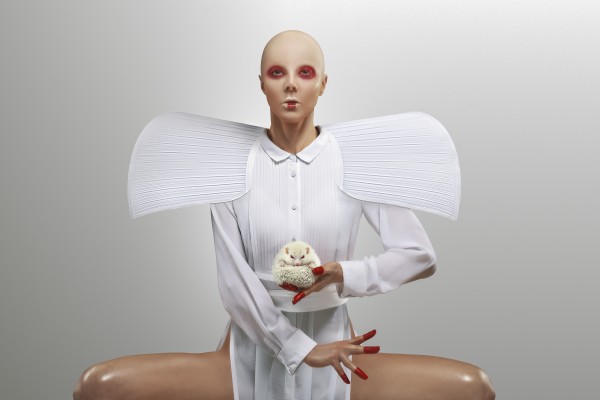 Front view of a bald woman wearing a white dress with large shoulder pads and red makeup holding an albino hedgehog standing in a white space by Artmood Visualz